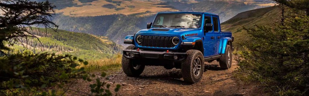 A blue 2025 Jeep Gladiator navigating rocky, mountainous terrain near Princeton, IL, with lush green and brown mountain range views and tree foliage in the foreground.