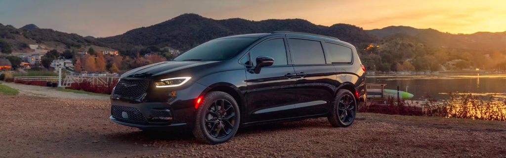 A black 2025 Chrysler Pacifica with headlights on, parked by a lake at sunset, with tall hills, mountains, and homes silhouetted against the glowing sky near Princeton, IL.