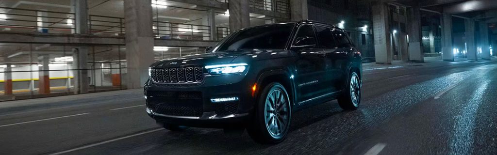 A black 2025 Jeep Grand Cherokee with headlights on, cruising through city streets at night near Princeton CDJR.