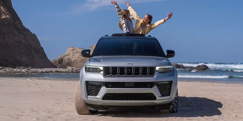 A silver Jeep Grand Cherokee parked on a beach with two people playfully posing on the roof under a clear blue sky.