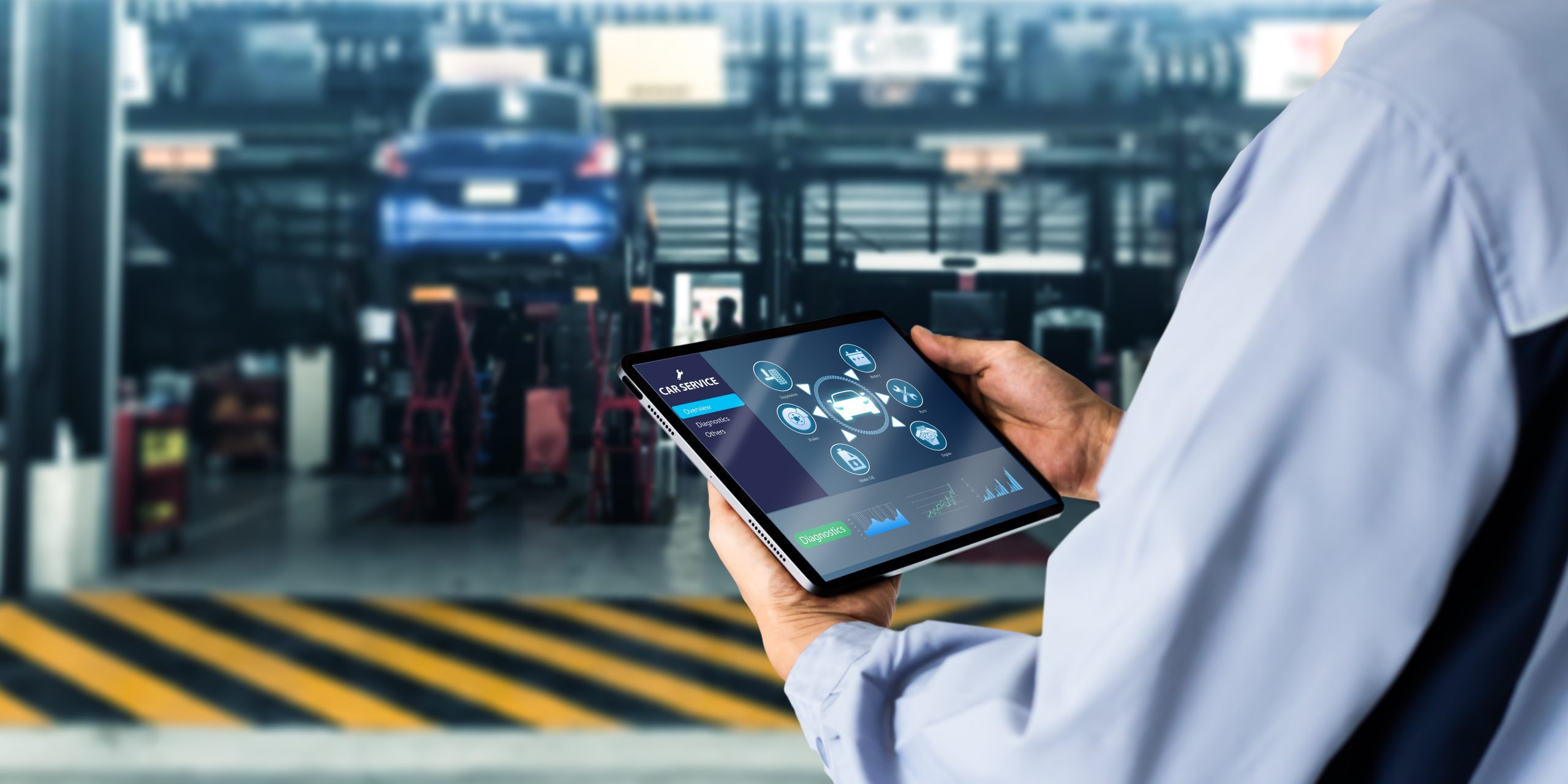 A technician holding a tablet displaying car service diagnostics in an automotive workshop.