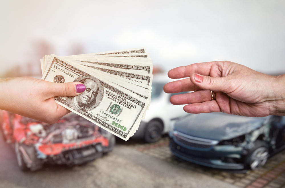 A hand with cash is reaching out to another hand against a backdrop of damaged cars.